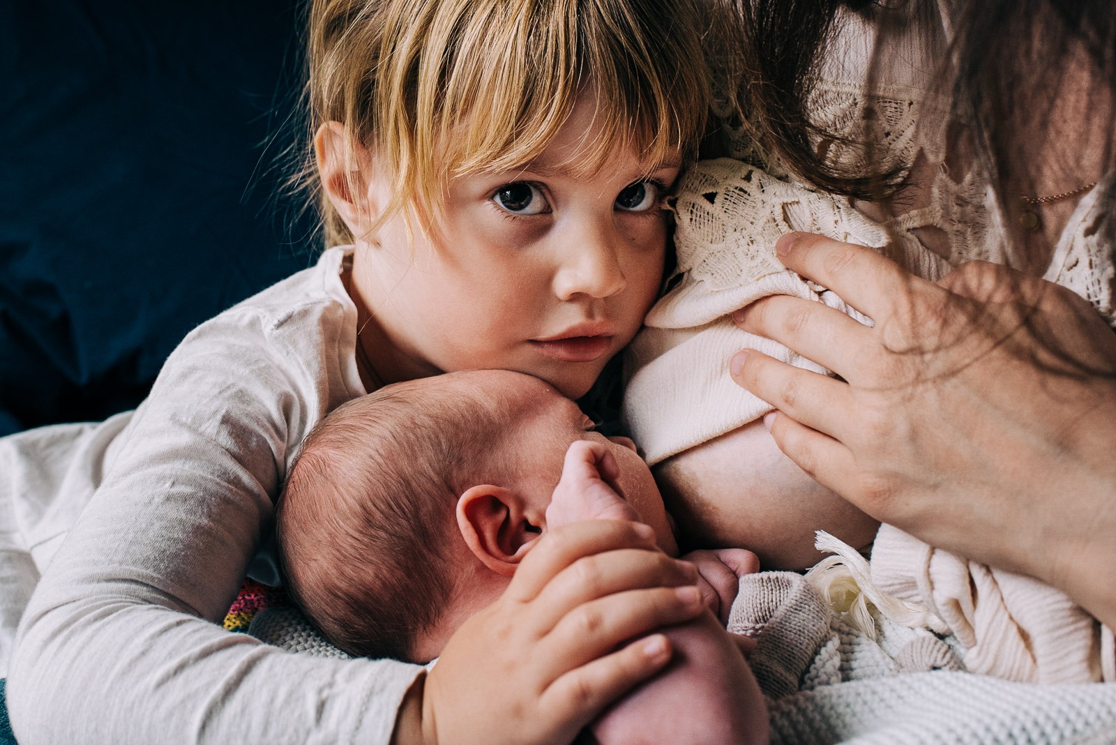 Eine Mutter stillt ihr Neugeborenes, die ältere Tochter hält ihr Geschwisterchen.
