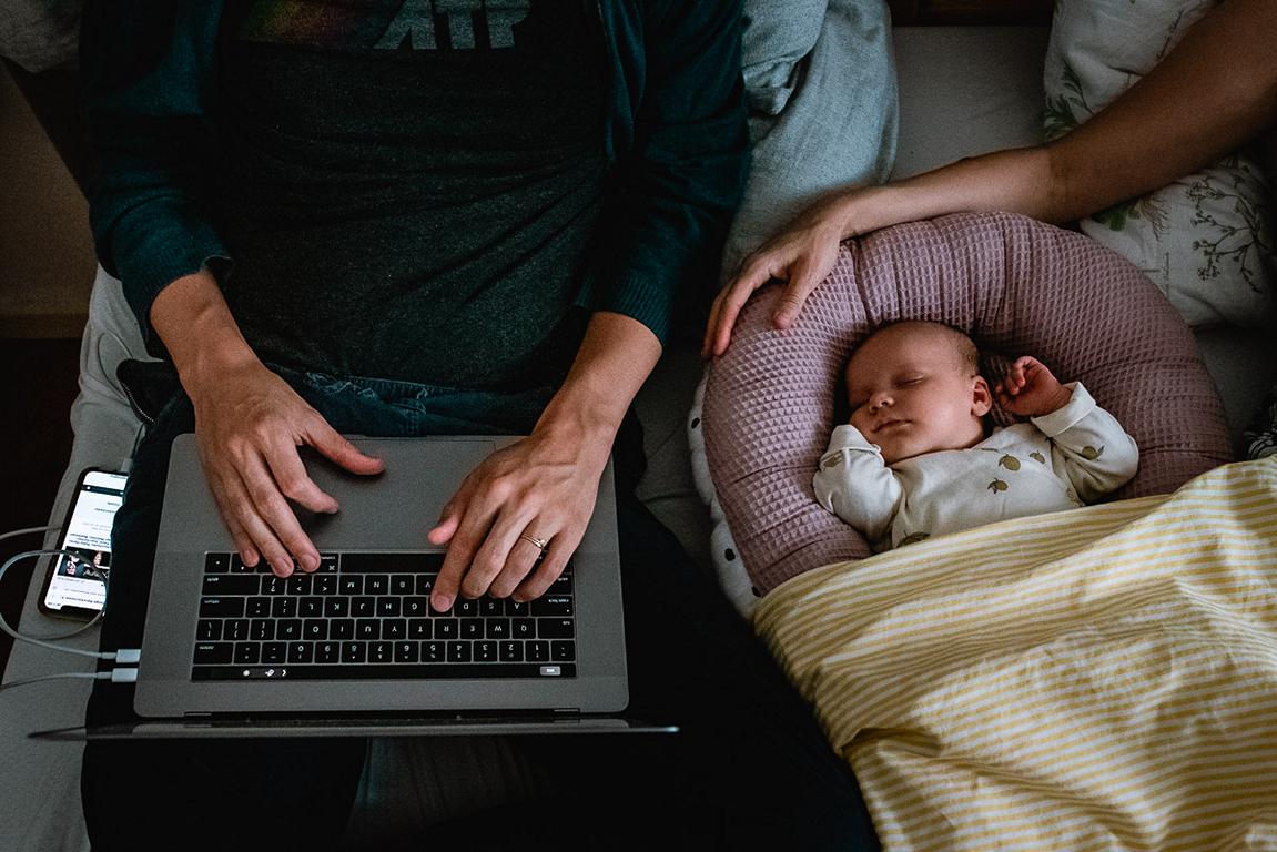 Der Blick von oben. Zwei Hände liegen auf einer Laptop-Tastatur. Daneben liegend ein schlafendes Baby. Der Arm der Mutter umarmt das Babynestchen.