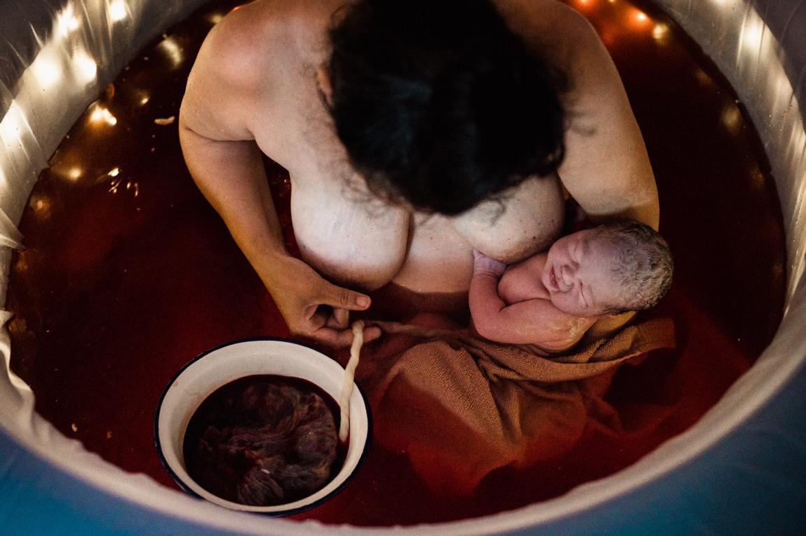 Blick von oben auf eine Mutter im Geburtspool, sie hält ihr Neugeborenes im Arm.