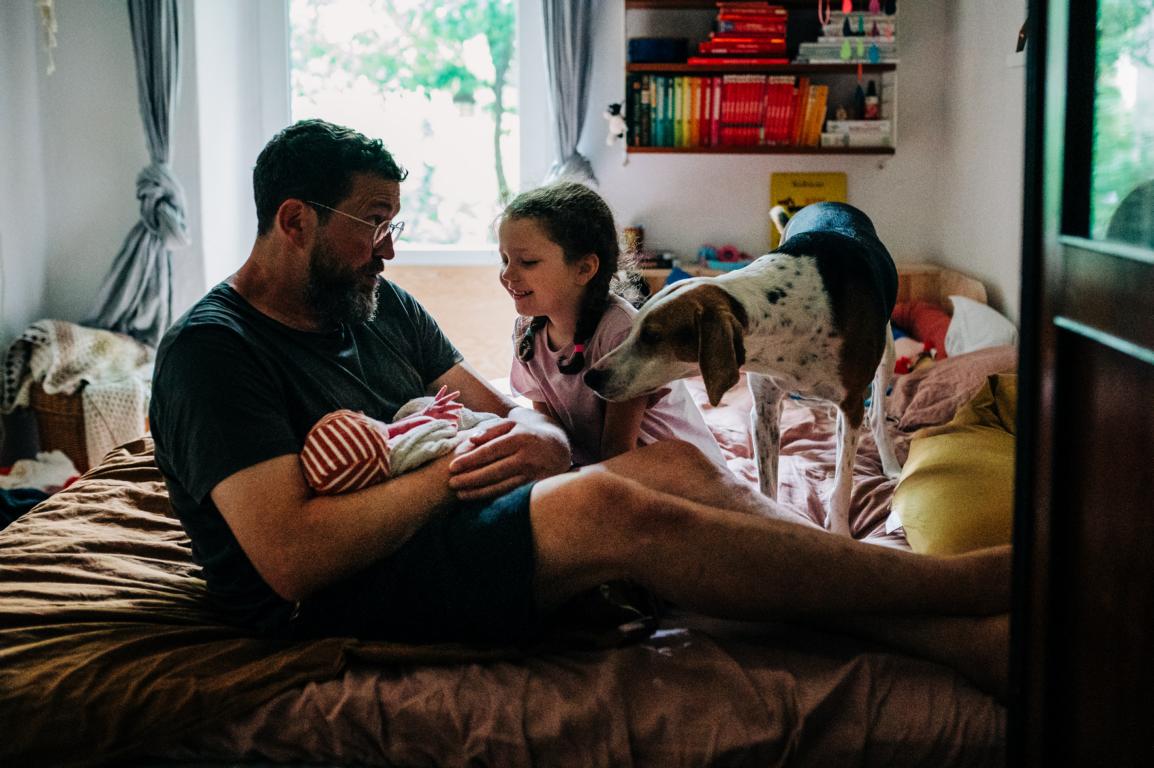 Der Vater hält glücklich sein Neugeborenes im Arm, neben ihm die ältere Tochter und der Familienhund.