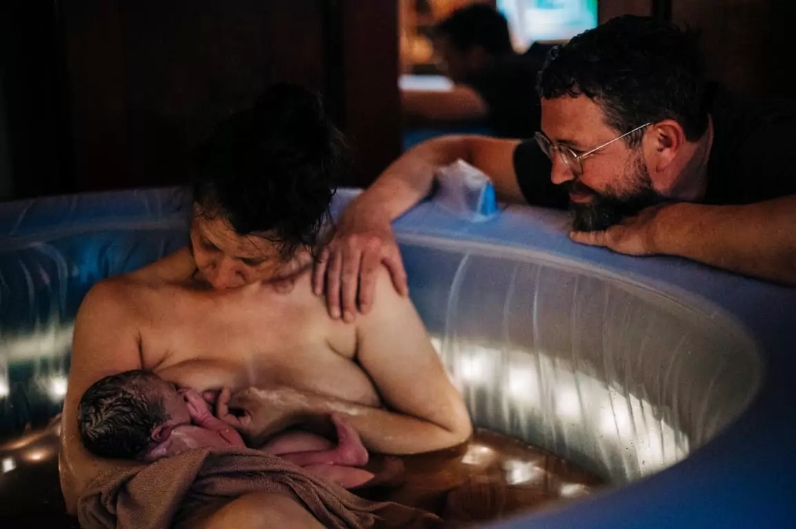 Eine Mutter sitzt im Geburtspool und stillt das Kind, der Vater beobachtet sie glücklich.