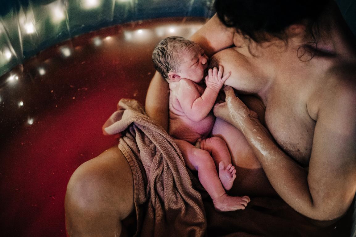 Eine eindrucksvolle Aufnahme der Geburtsfotografie: Das erste Stillen kurz nach der Geburt. Mutter und Baby im Geburtspool.