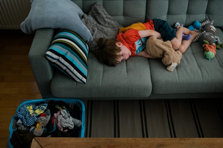 Ein Junge liegt erschöpft mit seinem Kuscheltier auf der Couch, neben ihm der volle Wäschekorb.
