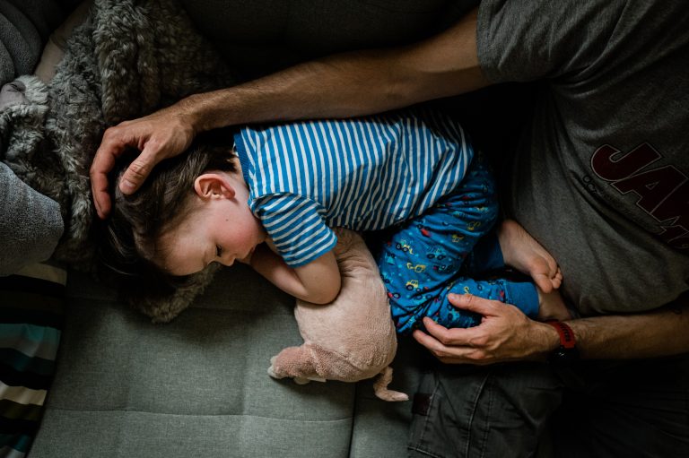 Ein Junge schläft mit seinem Kuscheltier auf der Couch, sein Vater hält ihn im Arm.